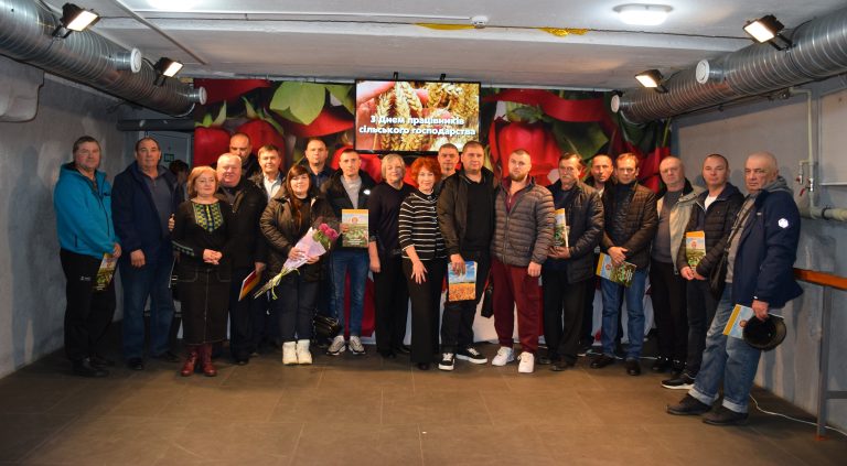 Кілька людей стоять у приміщенні перед великим муралом із зображенням овочів і монітором із написом «З Днем працівників сільського господарства».