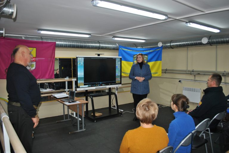 Жінка виступає перед аудиторією в приміщенні з великим телевізором та прапорами на тлі.