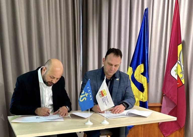 Два чоловіки в піджаках сидять по обидва боки столу, підписують документи між прапорами із символами.