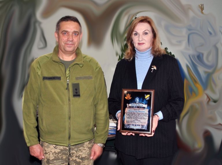 Жінка у чорному піджаку та світло-блакитному светрі тримає рамку з написом 