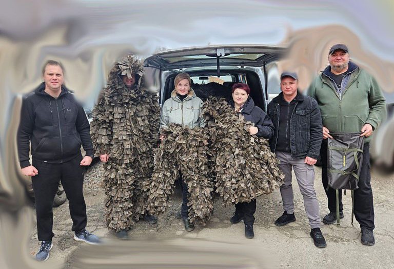 Чоловік та жінка у камуфляжних костюмах стоять поруч із трьома людьми у звичайному одязі біля автомобіля.