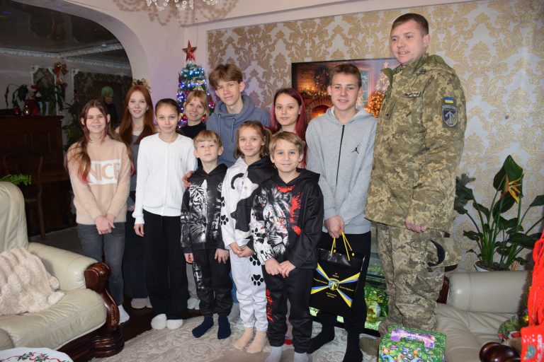 Військовий у камуфляжній формі стоїть поряд із дітьми в кімнаті, прикрашеній новорічними прикрасами та ялинкою.