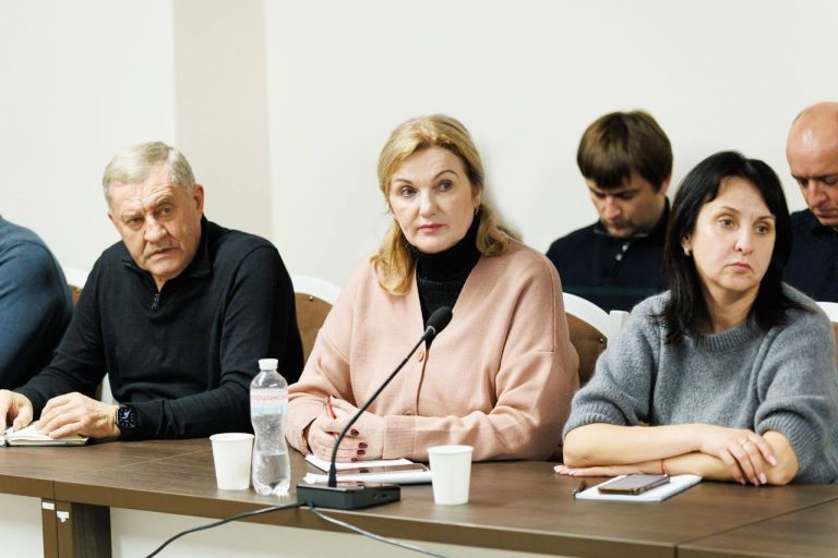 Жінка у рожевому та чорному одяді сидить біля мікрофона, облокотячись на стіл та тримаючи руки біля зошита і ручки.