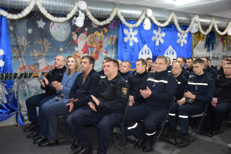 Чоловіки та жінки в офіційному одязі сидять і аплодують у приміщенні, прикрашеному зимовими святами.