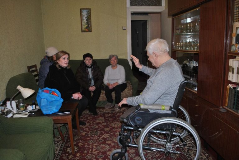 Літній чоловік у візку розмовляє з жінками, що сидять на дивані.
