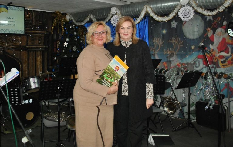 Жінка у світ-коричневому вбранні тримає книжку, поряд інша жінка у темному одязі.