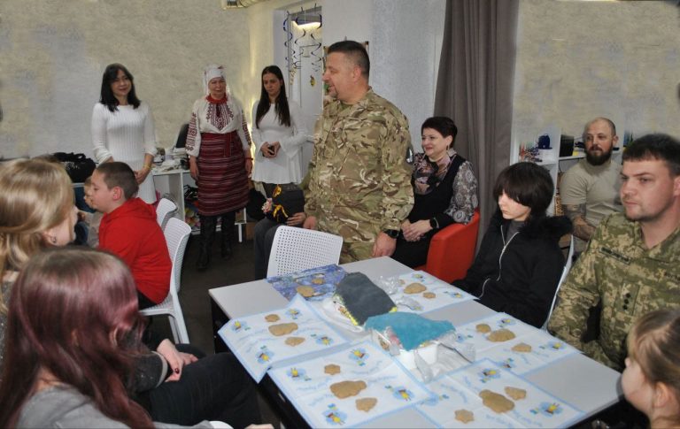 Чоловік у військовій формі стоїть біля столу, на якому лежать дитячі розмальовки та інші матеріали, серед дітей і жінок.
