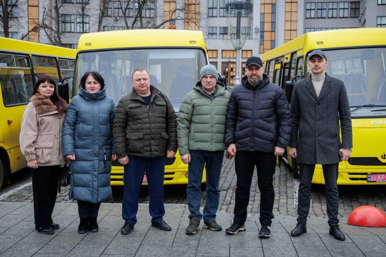 Шість людей стоять перед двома жовтими автобусами, серед яких жінки у пальто та чоловіки у пуховиках і шапках.