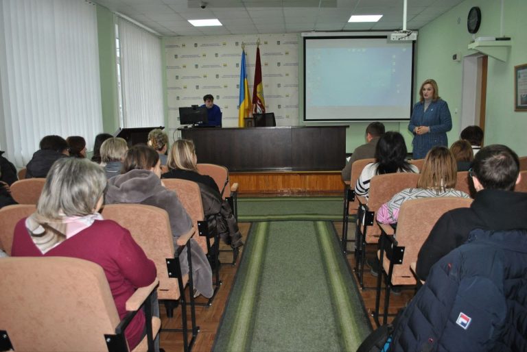 Жінка виступає перед аудиторією в офіційній обстановці, біля стіни із українським прапором та екраном для презентацій.