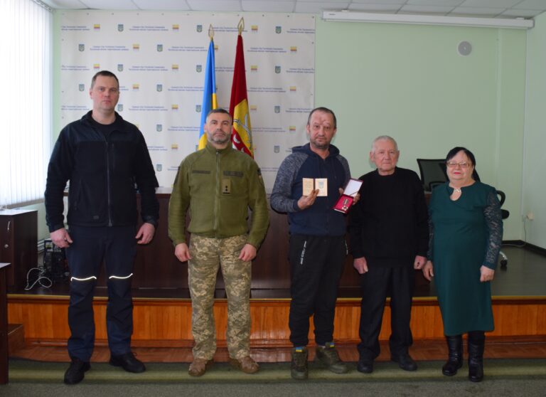 Чоловік у темно-сіній художці тримає в руках документи та нагороду.