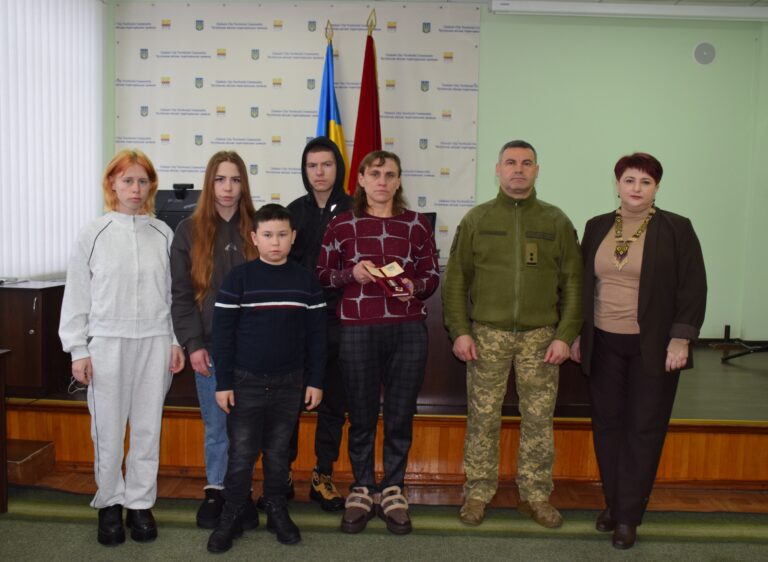 Жінка у червоному тримає нагороду, поряд стоять діти і чоловік у військовій формі і жінка у чорному піджаку.