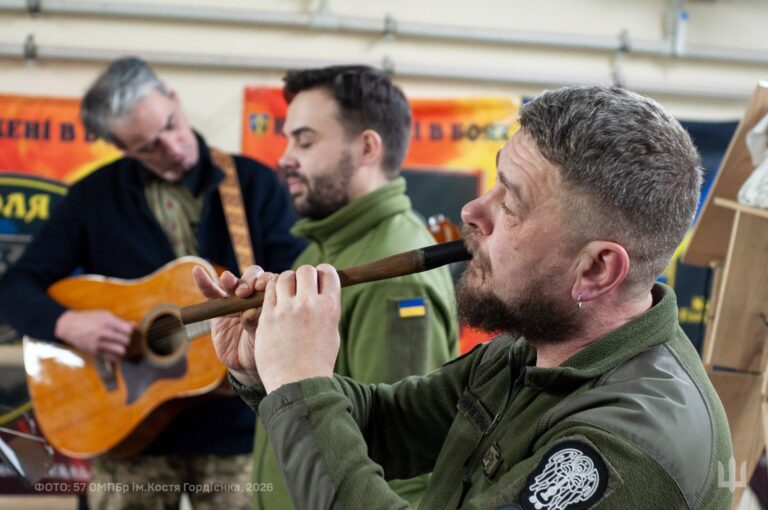 Чоловік у військовій формі грає на сопілі, інші музиканти на задньому плані грають на гітарі.