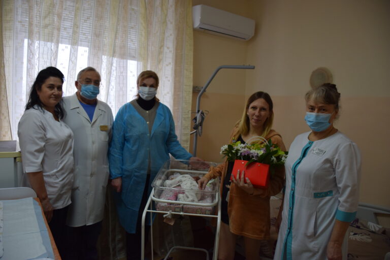 Жінка в помаранчевому одязі тримає квіти та немовля в ліжечку, оточена чотирма медичними працівниками у білих та синіх халатах і масках.