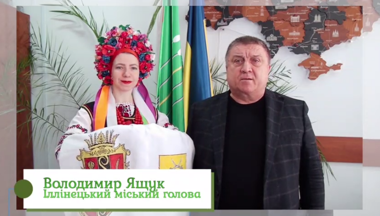 Чоловік у темному піджаку та жінка у вишитому вбранні з квітчастю на голові стоять поруч із прапорами та ілюстрацією української мапи.