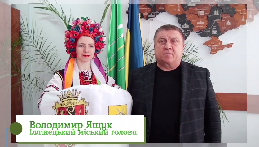 Чоловік у темному піджаку та жінка у вишитому вбранні з квітчастю на голові стоять поруч із прапорами та ілюстрацією української мапи.