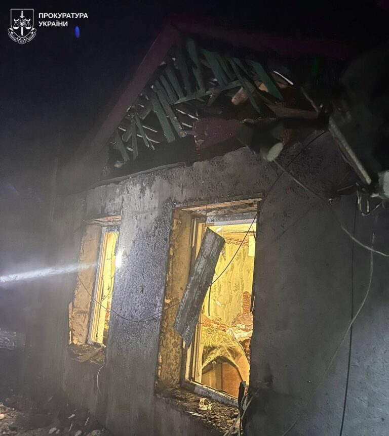 Будинок з пошкодженою стіною та вибитими вікнами, освітлений ізсередини, із димом та слідами вогню.