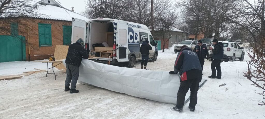 Чоловіки в зимньому одязі завантажують великий згорнутий білий матрас до вантажного автомобіля.