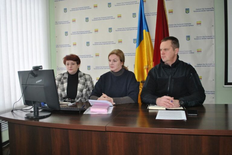 Жінка та двоє чоловіків сидять за столом із комп'ютером і документами, перебуваючи в офіційному приміщенні з українським прапором.