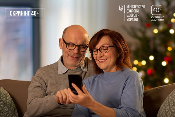 Літній чоловік та жінка сидять на дивані і разом дивляться на смартфон, посміхаючись, на тлі різдвяної ялинки із вогниками.