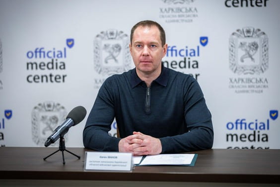 Чоловік сидить за столом із мікрофоном, склавши руки разом, перед ним лежить табличка з ім'ям та папка з документами.