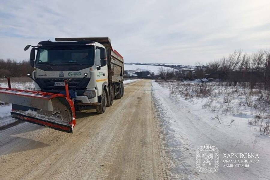 Велика біла вантажівка з снігоприбиральною машиною на переду їде по сільській дорозі вкритою снігом.