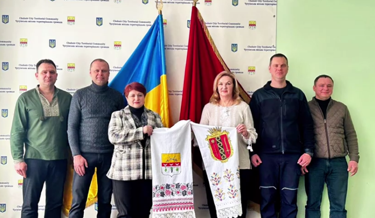 Шість людей різних статей та віку тримають велике вишиване рушник із зображенням герба на тлі прапорів України та Черкаської територіальної громади.