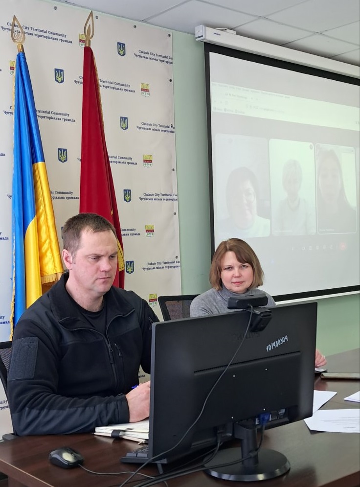 Чоловік і жінка сидять у конференц-залі за столом із комп'ютером, уважно слухаючи презентацію на великому екрані.