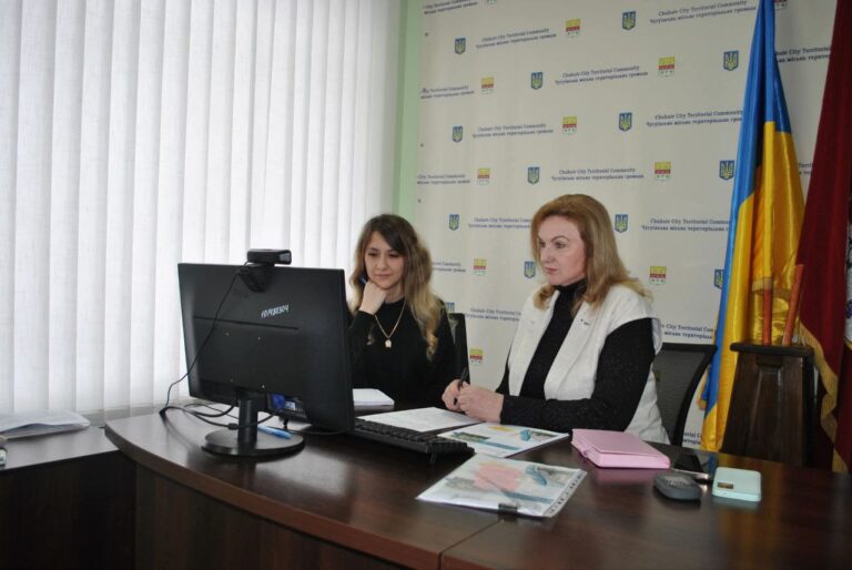 Дві жінки сидять за столом із комп'ютером і документами, розглядаючи інформацію перед офіційним фоном із символами територіальної громади.