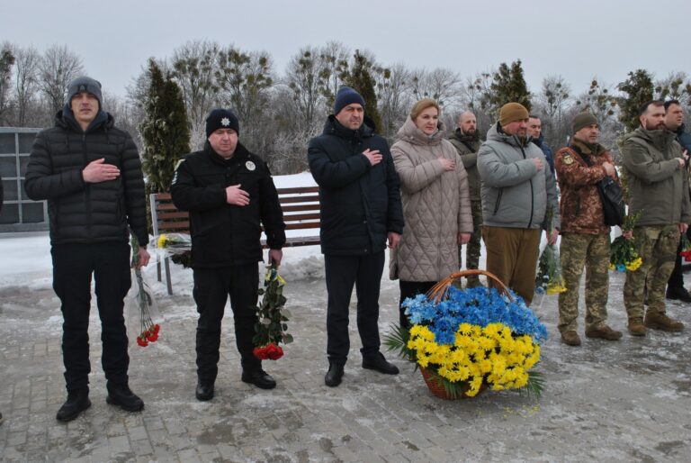 Група людей у зимовому одязі стоїть на вулиці, поклавши руку на серце й тримаючи квіти, біля кошика із жовто-блакитними квітами.