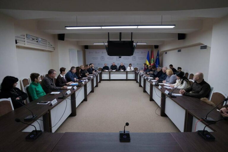Кімната засідань з довгими столами, мікрофонами та учасниками, розташованими по обидва боки, із трибуною на підвищенні в дальньому кінці кімнати.
