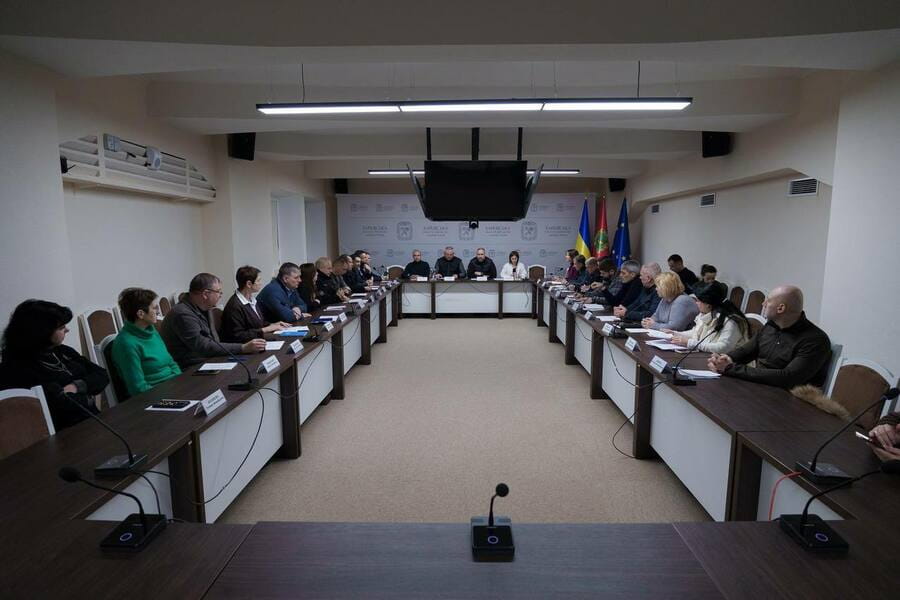 Кімната засідань з довгими столами, мікрофонами та учасниками, розташованими по обидва боки, із трибуною на підвищенні в дальньому кінці кімнати.