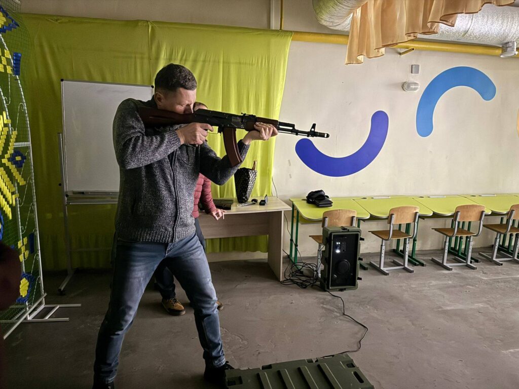 Чоловік у светри та джинсах цілиться з автомата у приміщенні класного кімнати зі світлою стіною та лінійкою парт.