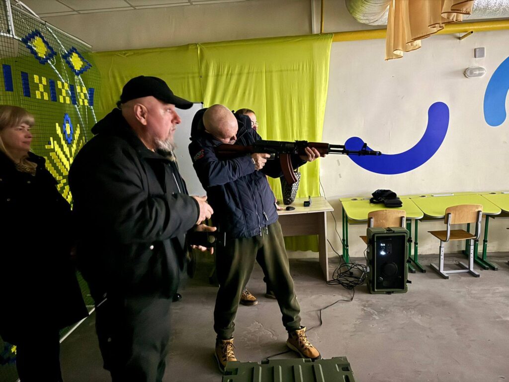 Чоловік у темній куртці та капелюху навчає іншого чоловіка стріляти з автомата, третій чоловік стоїть позаду них.