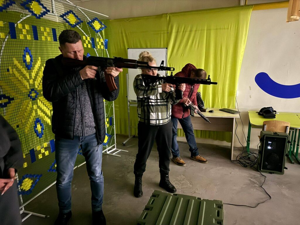Чоловік у темному одязі цілиться з автомата у приміщенні зі стіною жовтого кольору та інструктором у світлому одязі і жінкою у червоно-сірому одязі на задньому плані.