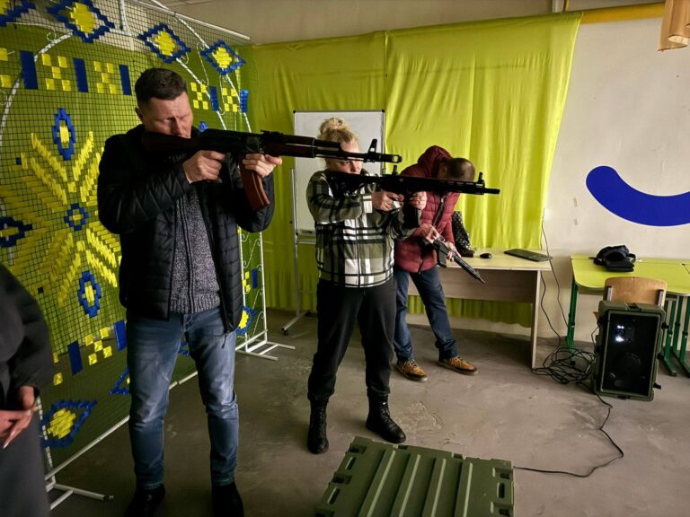 Чоловік у темному одязі цілиться з автомата у приміщенні зі стіною жовтого кольору та інструктором у світлому одязі і жінкою у червоно-сірому одязі на задньому плані.