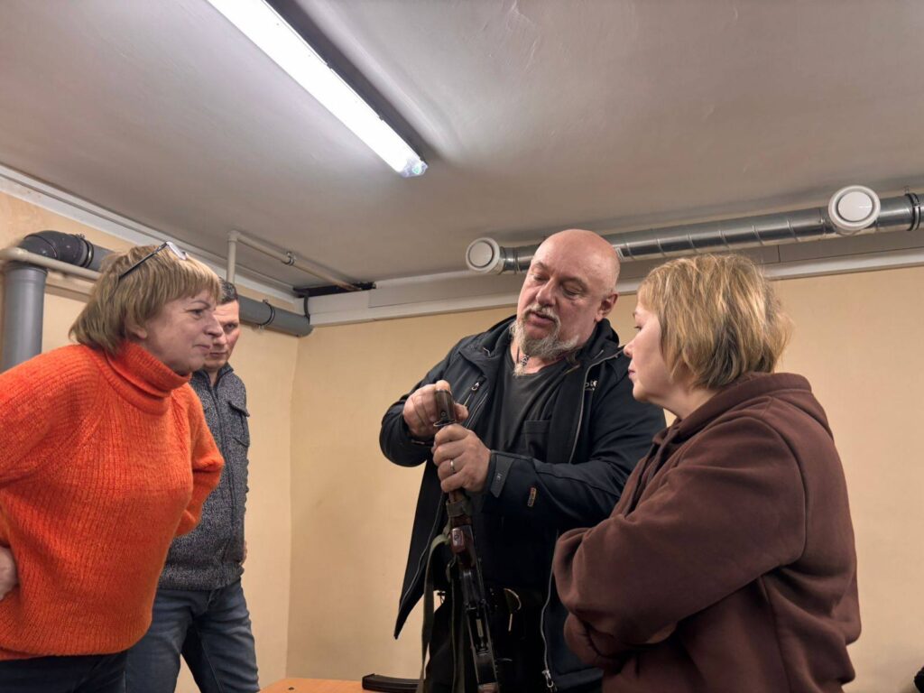 Чоловік демонструє жінкам пістолет, стоячи в приміщенні з жовтими стінами та трубами на стелі.