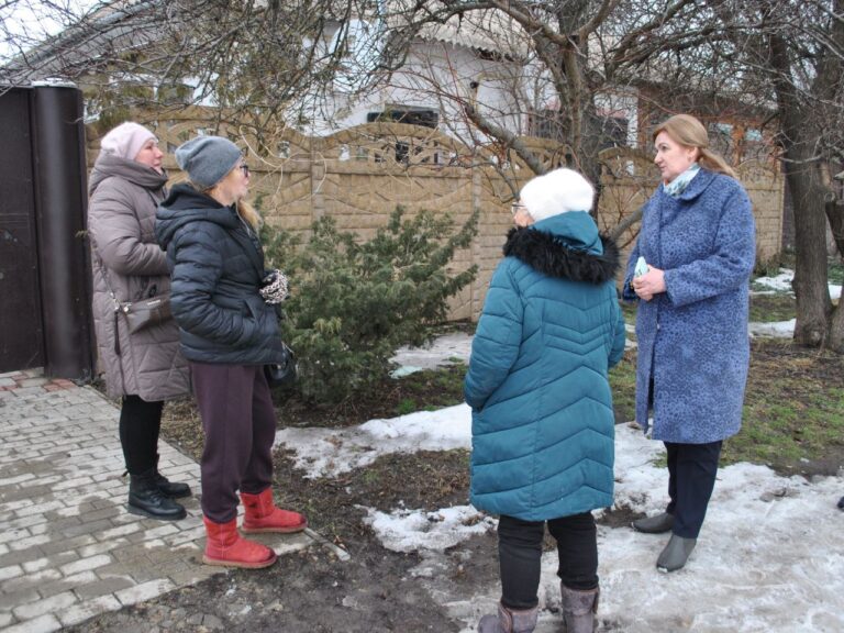 Чотири жінки в теплих зимових речах стоять і розмовляють біля будинку з цегляним забором та голими деревами.