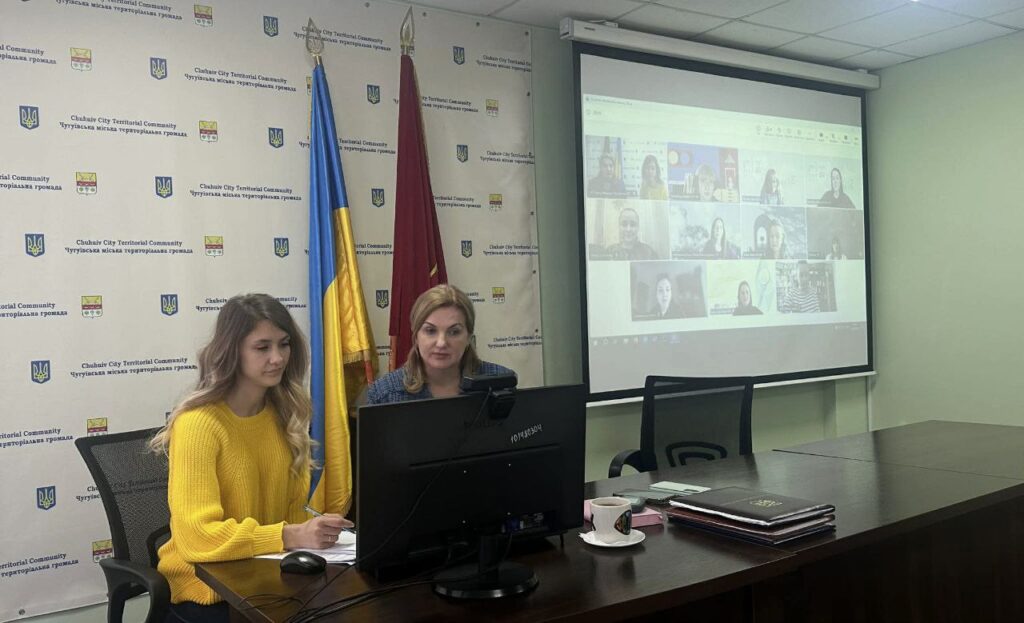 Дві жінки сидять перед монітором комп'ютера та беруть участь у відеоконференції, представленій на великому екрані.
