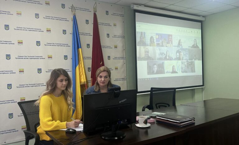 Дві жінки сидять перед монітором комп'ютера та беруть участь у відеоконференції, представленій на великому екрані.