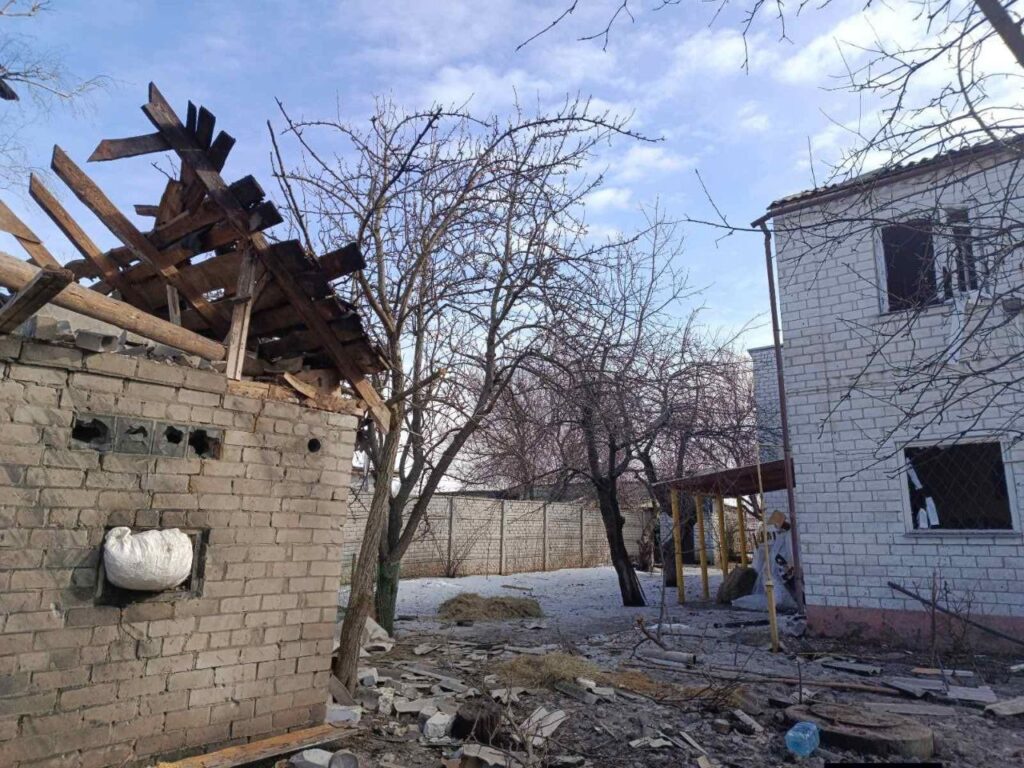 Пошкоджена будівля з частково зруйнованим дахом та вибитими вікнами, поруч стоїть непошкоджений будинок із закритими вікнами та дверима.