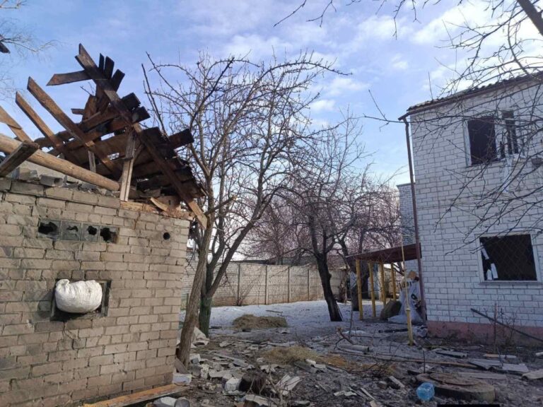 Пошкоджена будівля з частково зруйнованим дахом та вибитими вікнами, поруч стоїть непошкоджений будинок із закритими вікнами та дверима.