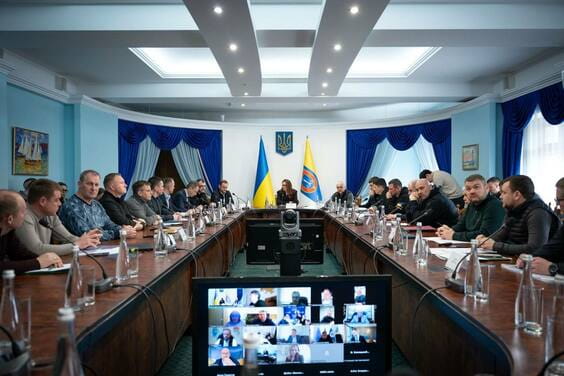Жінка веде нараду в офіційній кімнаті з українським прапором та іншими учасниками, деякі з яких підключені до відеозв'язку.