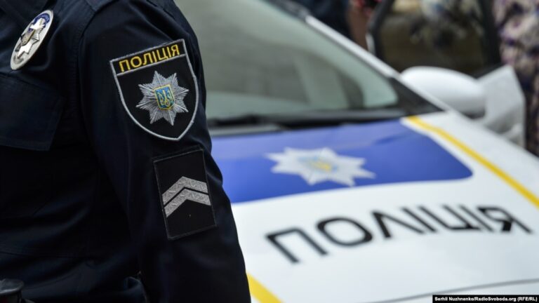 Поліцейський у формі з нашивками правоохоронної служби поряд із поліцейським автомобілем.
