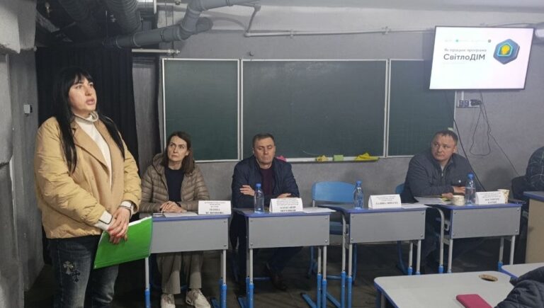 Жінка стоїть біля столу, на якому сидять троє людей і іще один учасник частково видно праворуч.