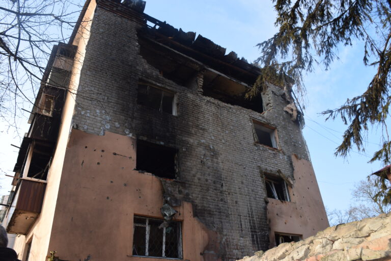 Пошкоджена будівля з чорними слідами від вогню та вирвами від куль чи вибухів на стінах.