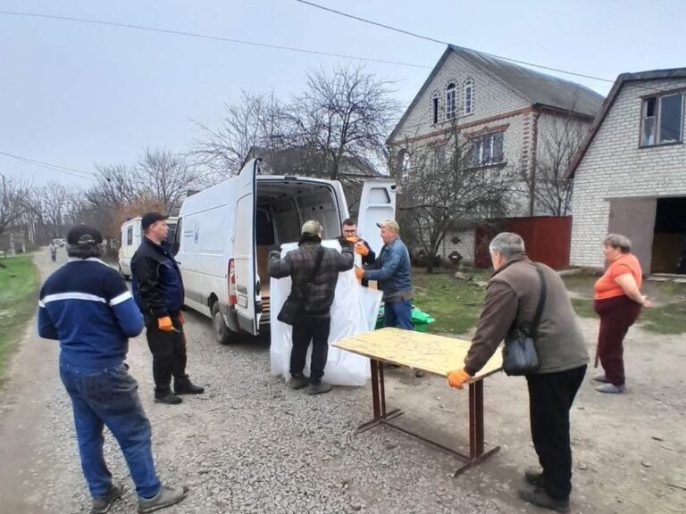Чоловіки розвантажують білі мішки з білого фургона, що стоїть біля будинку, а інші люди стоять поруч і спостерігають.