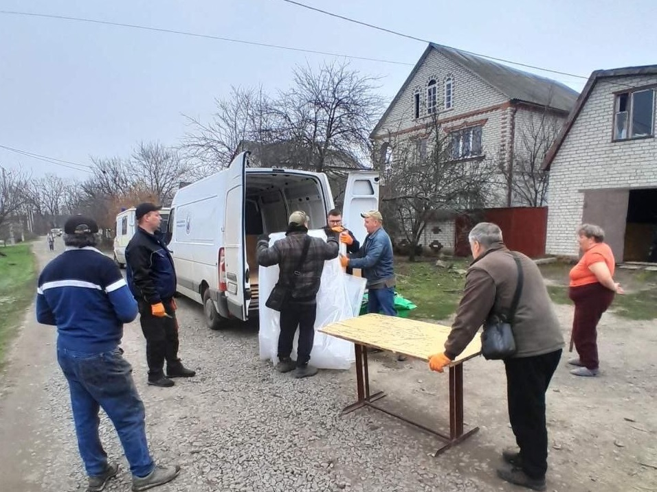 Чоловіки розвантажують білі мішки з білого фургона, що стоїть біля будинку, а інші люди стоять поруч і спостерігають.