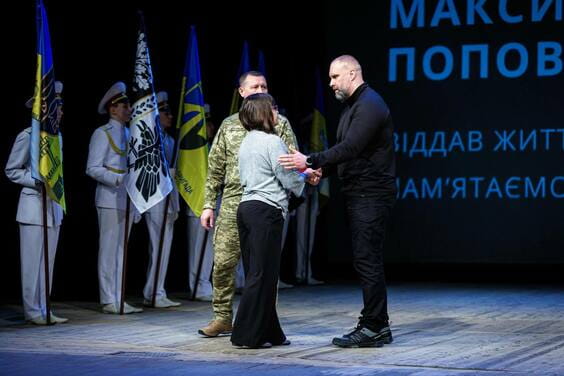Чоловік у військовій формі обіймає жінку, а чоловік у чорному одязі стоїть поруч і дивиться на них; позаду групи стоять військові із прапорами.