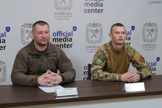 Два чоловіки в військовому одязі сидять за столом із табличками і паперами, на тлі баннера із написом 