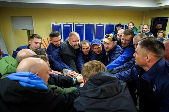 Чоловіки в синіх та чорних куртках зібрались у коло та склали руки разом, демонструючи єдність та підтримку один одного.